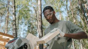 Man wearing safety gear cuts wood outdoors with power tools, emphasizing craftsmanship.