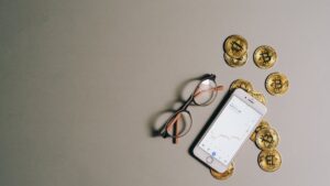 Flat lay of Bitcoin coins, smartphone with graph, and glasses on gray surface.
