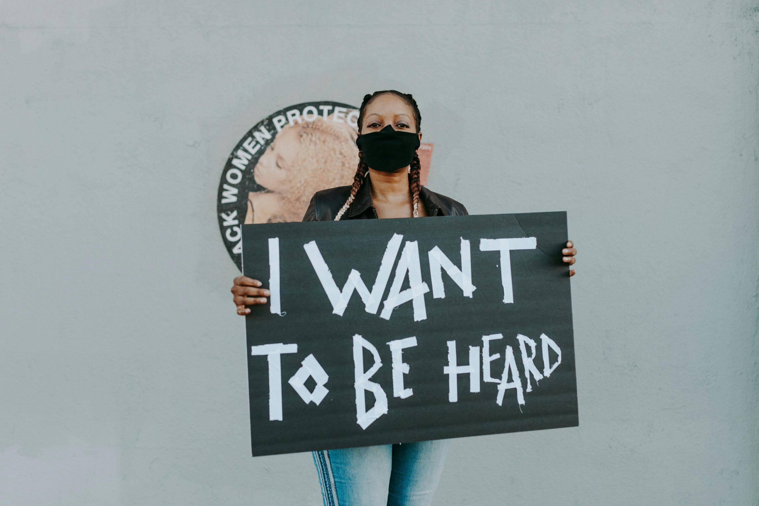 Masked protester holds a sign saying 'I Want to Be Heard' against a wall with activist imagery.