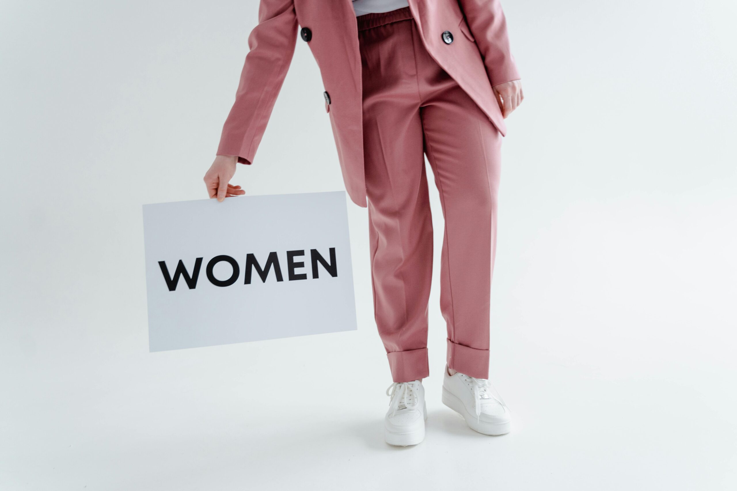 Close-up of a woman in a pink suit holding a 'WOMEN' sign on a white background